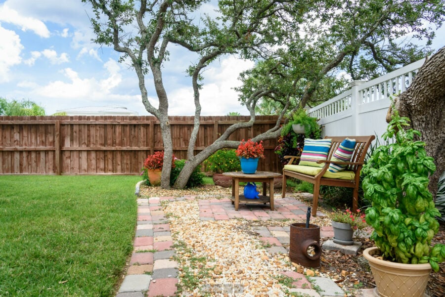 Cozy backyard garden with a mature tree, colorful potted flowers, brick pathway, and outdoor seating area featuring striped cushions, surrounded by a wooden privacy fence under a partly cloudy sky.