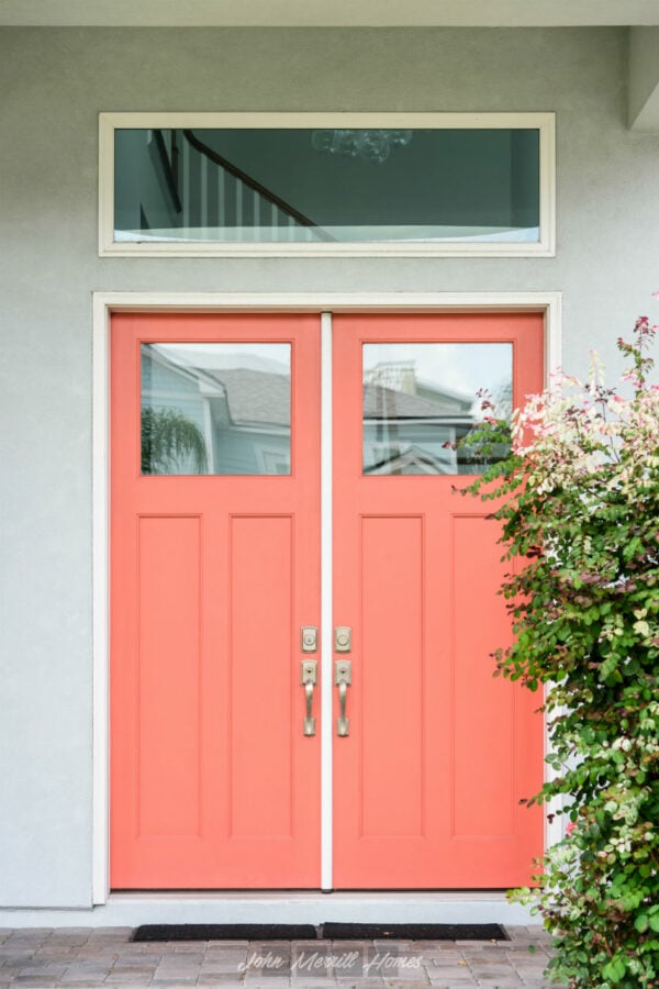 Beautiful exterior front door in coral color with decorative glass panels and modern hardware, surrounded by a light-colored house facade and lush greenery to enhance curb appeal and home entryway style.