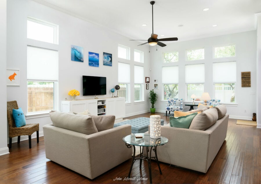 Bright and spacious living room with white walls, high ceilings, large windows with blinds, a modern ceiling fan, and stylish furniture and decor for a welcoming atmosphere.