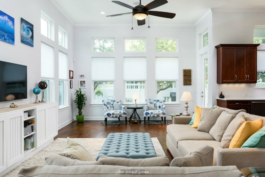 Bright, airy living room with large windows, white walls, and contemporary decor including a beige sofa with colorful pillows, a tufted ottoman, and a cozy seating area with floral-patterned chairs, creating a welcoming space.