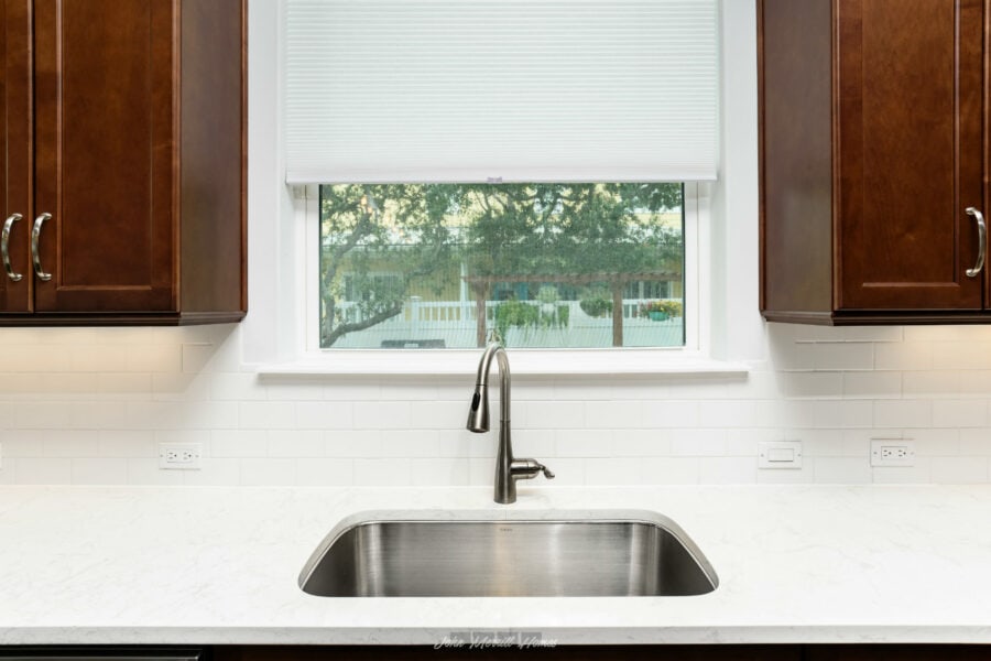 Modern kitchen sink with stainless steel faucet beneath a large window, featuring white subway tile backsplash, surrounded by dark wood cabinets and multiple electrical outlets for convenience.