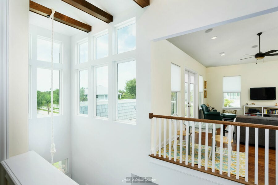 Bright second-floor view of a spacious living room with large windows, white walls, and wooden ceiling beams, featuring a comfortable seating area with a TV and colorful rug.