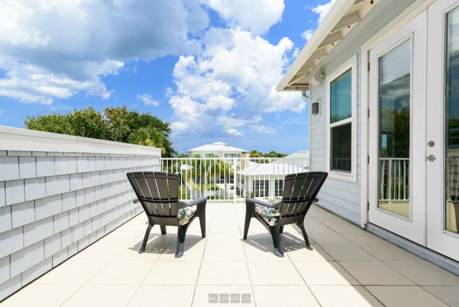 Modern white balcony with outdoor chairs, overlooking a vibrant neighborhood with greenery and a bright blue sky with scattered clouds.