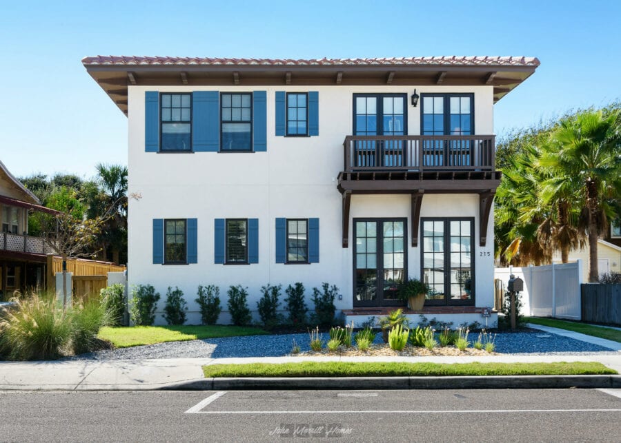 Modern coastal style house featuring white stucco exterior, dark blue window shutters, and a second-story balcony with wooden railing, surrounded by lush landscaping and palm trees.
