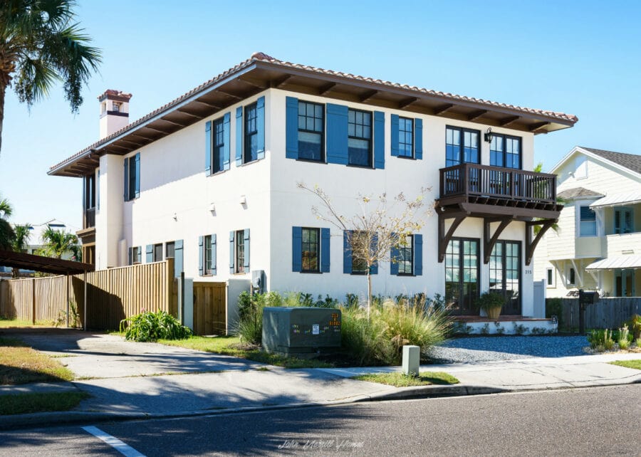 Modern white two-story home with blue window shutters, a small balcony, and landscaped yard in a sunny suburban neighborhood.