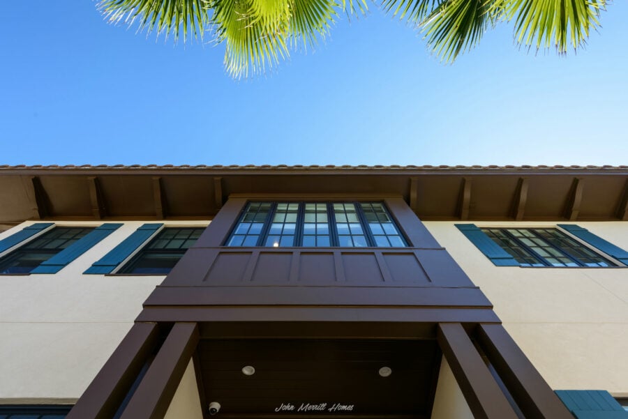 Modern two-story home exterior with large windows, dark brown accents, and a white facade, framed by a clear blue sky and lush green palm leaves.