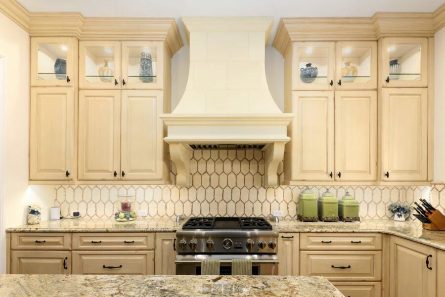 Warm cream-colored kitchen cabinets with black hardware, a matching beige tile backsplash, and a stainless steel stove with a decorative vent hood, creating a cozy and elegant cooking space.