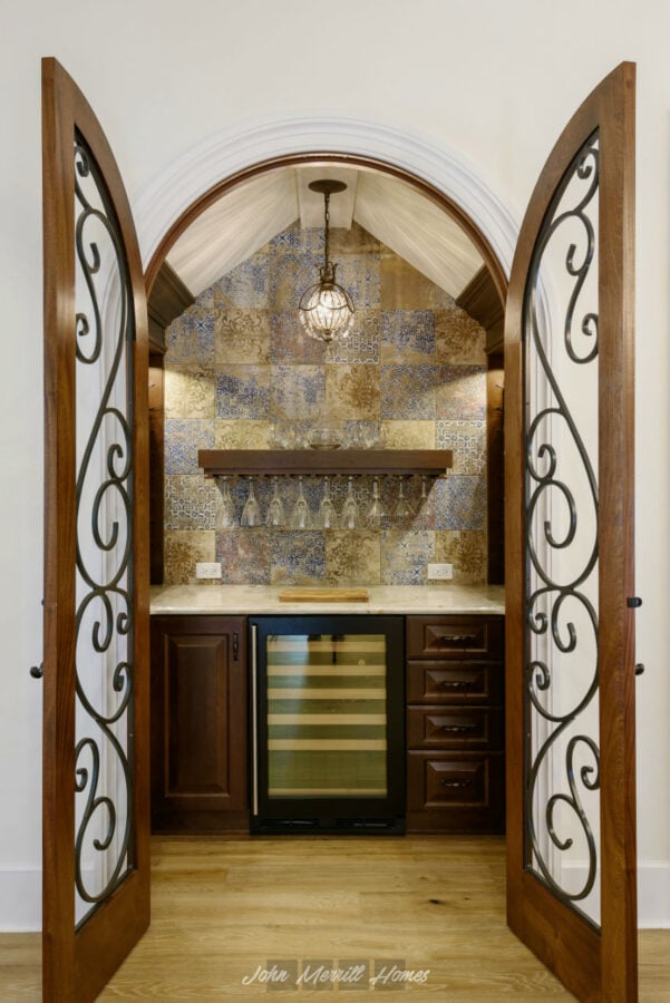 Warm wood and wrought iron double doors open to a stylish home wine bar with a built-in wine cooler, glass rack, and decorative backsplash tiles under a cozy hanging pendant light.