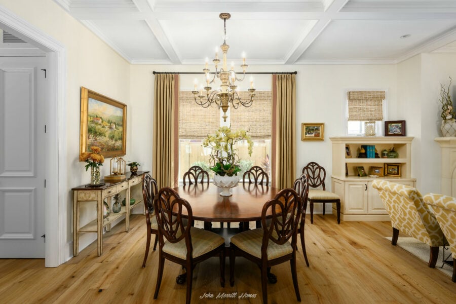Spacious dining room with a round wooden table, elegant chandelier, large windows with beige curtains, and classic furniture, creating a warm and inviting atmosphere.