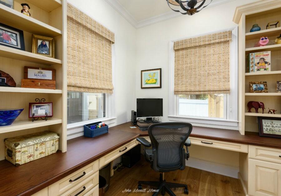 Cozy home office with built-in bookshelves, cream-colored cabinetry, and large windows with woven bamboo shades. A black ergonomic chair, desktop with office supplies, and framed artwork create a functional workspace.