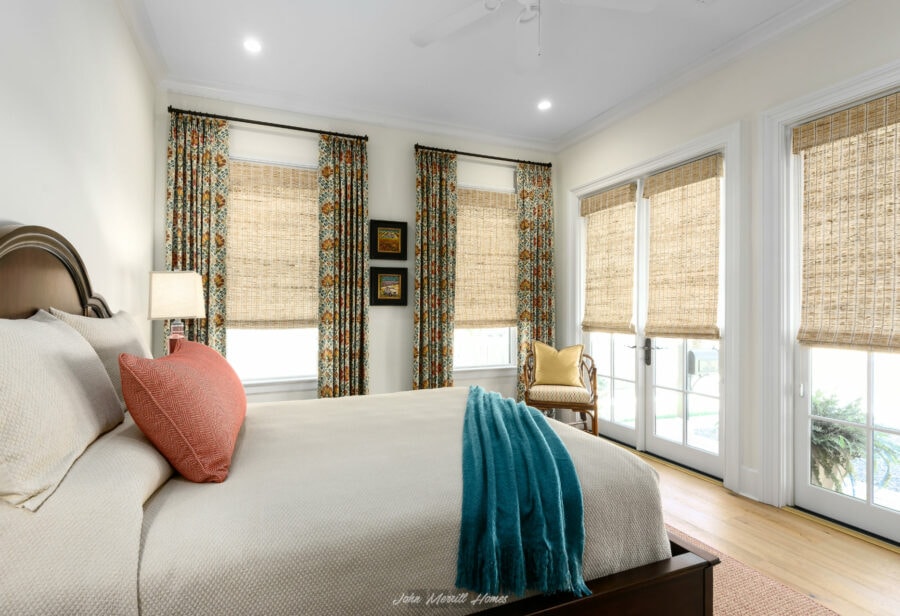 Bright and cozy bedroom with large windows covered by woven bamboo blinds and floral curtains, featuring a king-sized bed with colorful pillows, a wooden chair with a yellow pillow, and French doors leading outside.