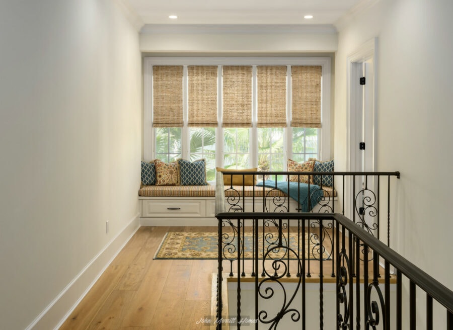 Cozy sitting nook with large multi-pane windows covered by woven bamboo shades, built-in window seat with decorative cushions, hardwood flooring, and ornate black iron stair railing.
