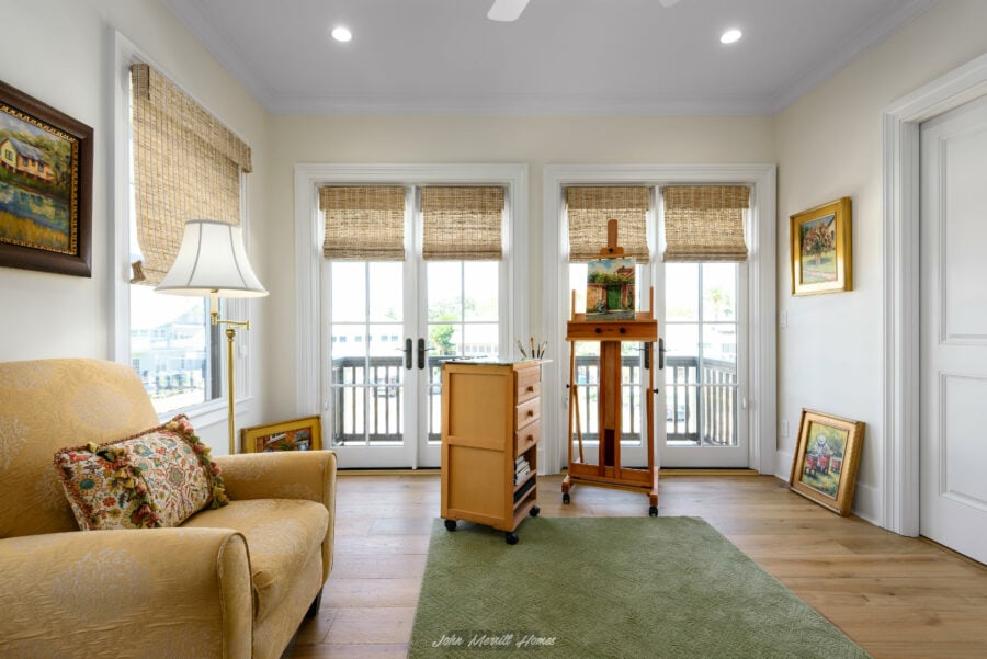 Bright living room with large windows, bamboo shades, yellow sofa, art easel, and wooden floors, creating a cozy and artistic space in a home built by John Merrill Homes.