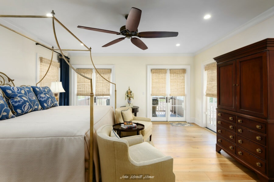 Luxurious master bedroom with a four-poster canopy bed, elegant armchair, and a wooden armoire, featuring woven blinds, a ceiling fan, and doors leading to an outdoor balcony.