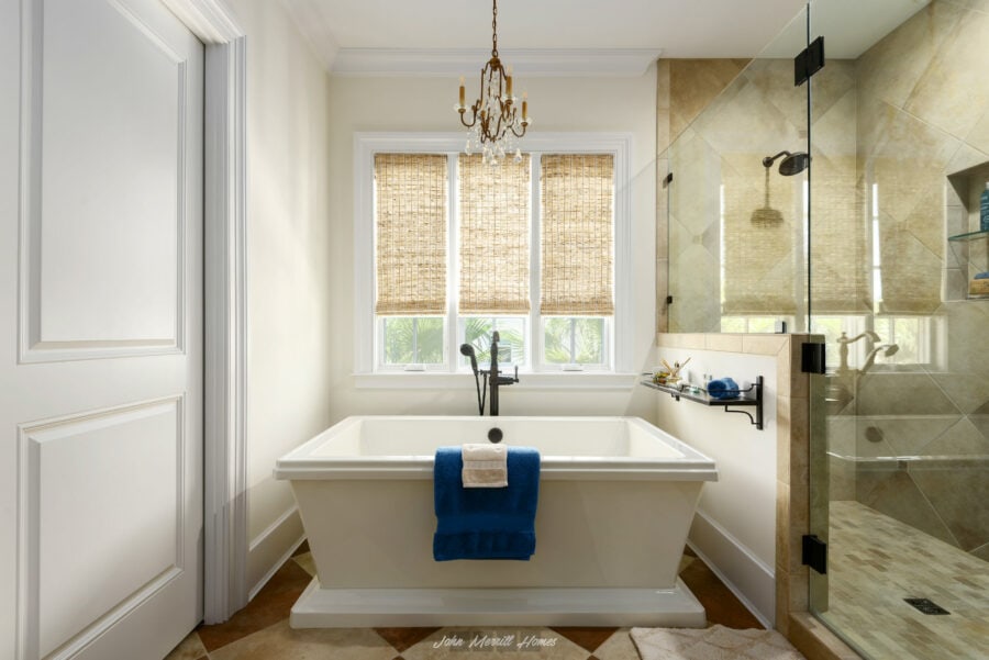 Luxurious bathroom featuring a white bathtub, large window with woven shades, elegant chandelier, and a glass-enclosed shower with beige tiles, creating a spa-like retreat.