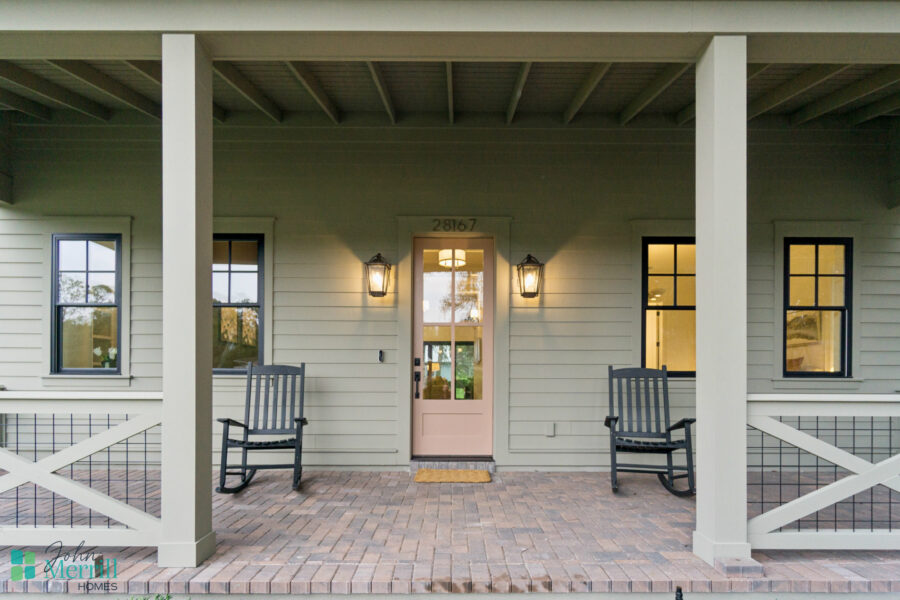 Elegant front porch of a modern house with two black rocking chairs, a pink front door, outdoor lighting fixtures, and a cozy welcoming atmosphere, showcasing home design by John Merrill Homes.