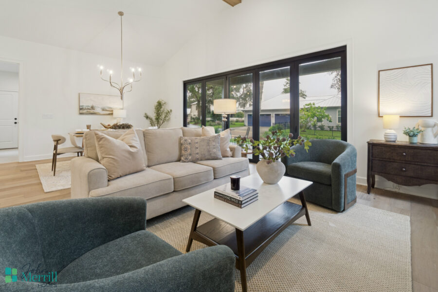 Bright and airy living room with modern decor, featuring a large beige sofa, accent chairs, and a coffee table, surrounded by large windows that showcase a lush outdoor backyard.