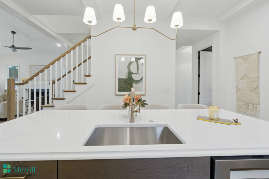 Bright modern kitchen with white marble countertops, stainless steel sink, and decorative flower arrangement. Open-concept design featuring a staircase, artwork, and soft lighting for a welcoming home environment.