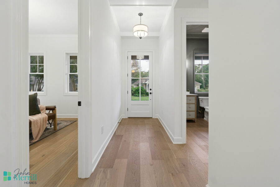 Bright white interior hallway with a front door, hardwood flooring, and natural light from adjacent rooms, showcasing modern home design by John Merrill Homes.