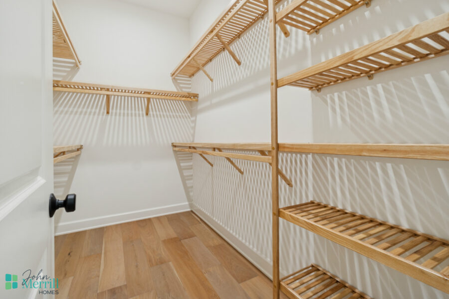Spacious walk-in closet with wooden shelves and hanging rods, bright white walls, and warm hardwood flooring, offering organized storage and natural light for a luxurious wardrobe space.
