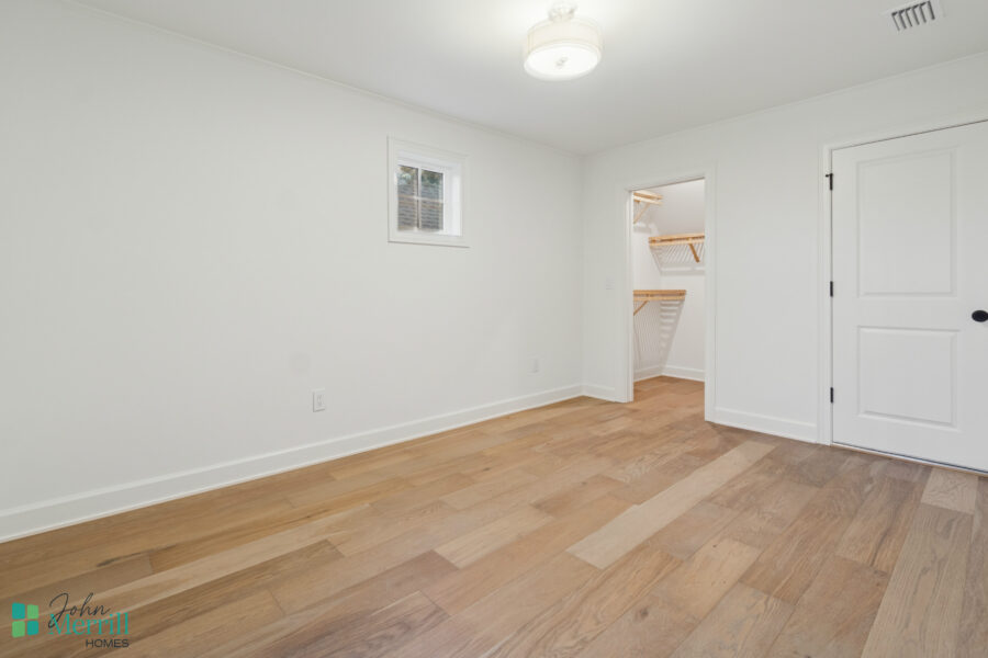 Bright and spacious interior room with white walls, small window, ceiling light fixture, hardwood flooring, and a walk-in closet with wooden shelving, part of a modern home designed by John Merrill Homes.