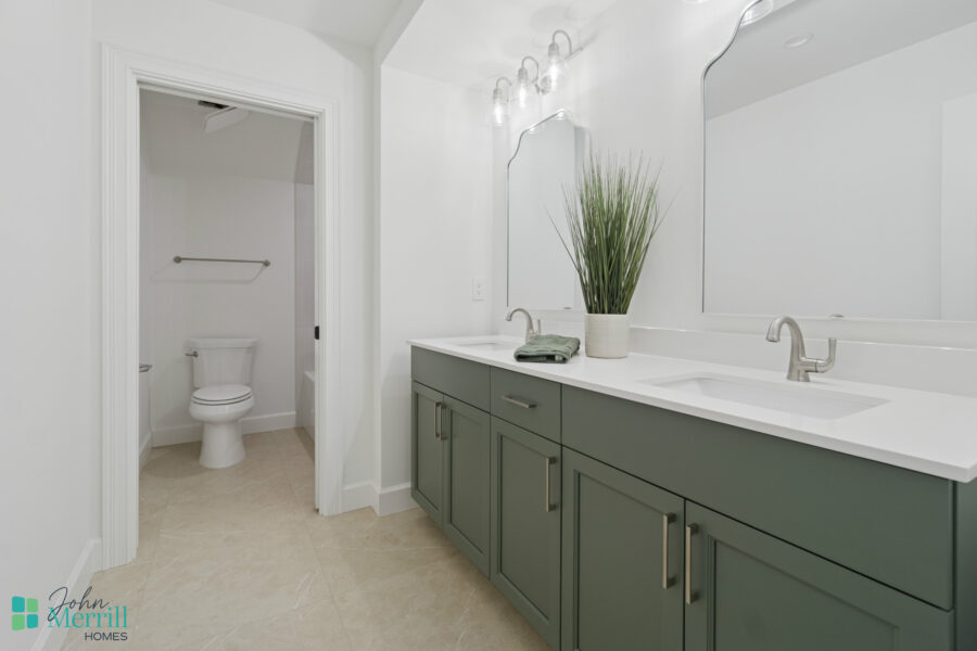 Bright modern bathroom with dual sinks, large mirror, fresh greenery, and neutral tile flooring; includes an adjacent toilet area for convenience.