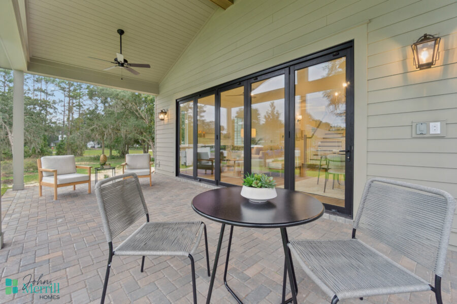 Bright covered outdoor patio with comfortable seating, potted plant, and large sliding glass doors leading to a spacious living area, set in a lush green backyard with tall trees.