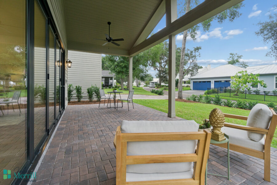 Spacious covered backyard patio with seating, potted decor, brick flooring, and outdoor ceiling fan, surrounded by lush greenery and residential neighborhood for relaxing outdoor living.