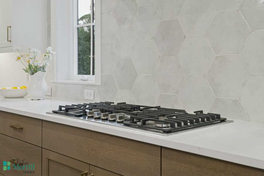 Modern kitchen with white countertop, built-in gas stove, and beige cabinetry, featuring a textured hexagon tile backsplash and a window with greenery view.