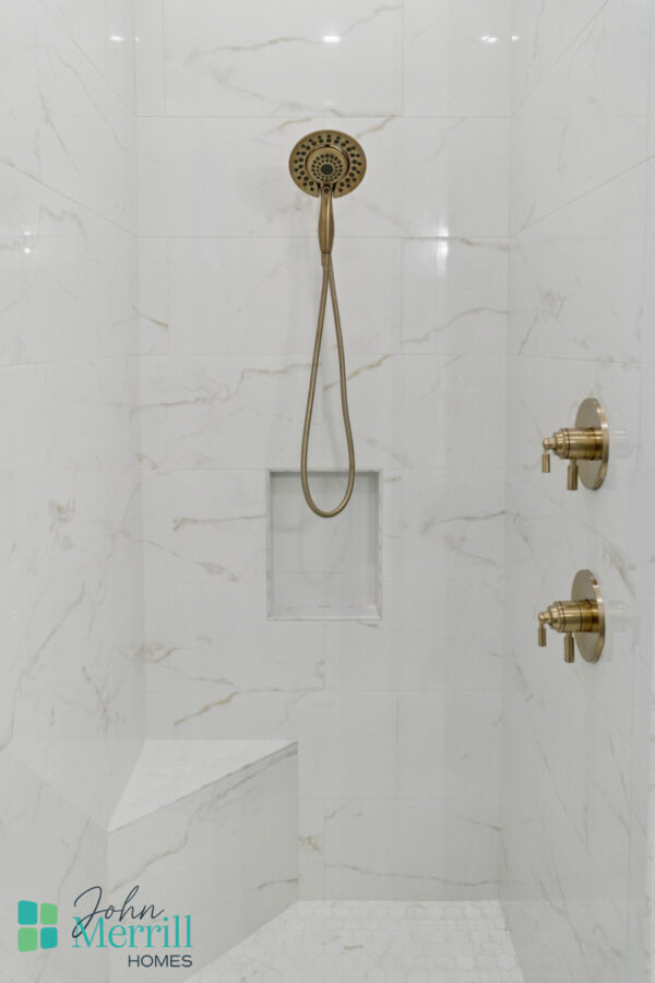 Luxurious walk-in shower with polished gold fixtures, marble-like wall tiles, built-in shelf, and a corner seat, showcasing modern bathroom design by John Merrill Homes.