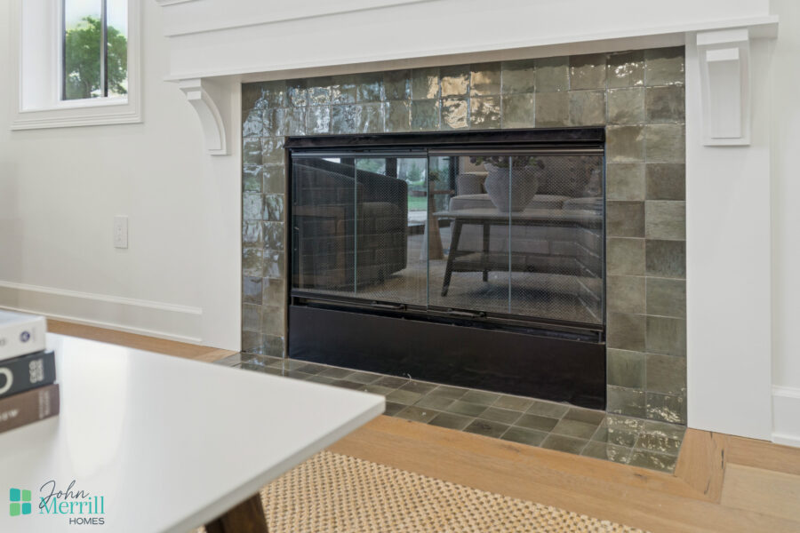 1. Modern living room fireplace with green and brown tiled surround, black metal screen, and white walls, featuring natural light from a nearby window and hardwood flooring.