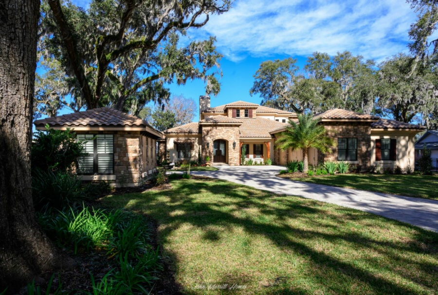 Luxury home with stone accents and a lush front yard, surrounded by mature trees under a bright blue sky, showcasing elegant landscaping and a welcoming entrance.