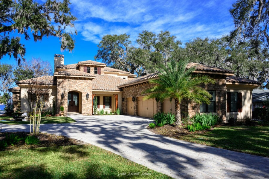 Luxurious single-family home with a stone facade, lush landscaping, and a paved driveway under a bright blue sky, showcasing elegant architecture and vibrant greenery.