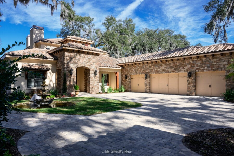 Luxurious single-story home with a stone and stucco exterior, three-car garage, Mediterranean-style clay tile roof, lush landscaping, and a picturesque fountain in the front yard.