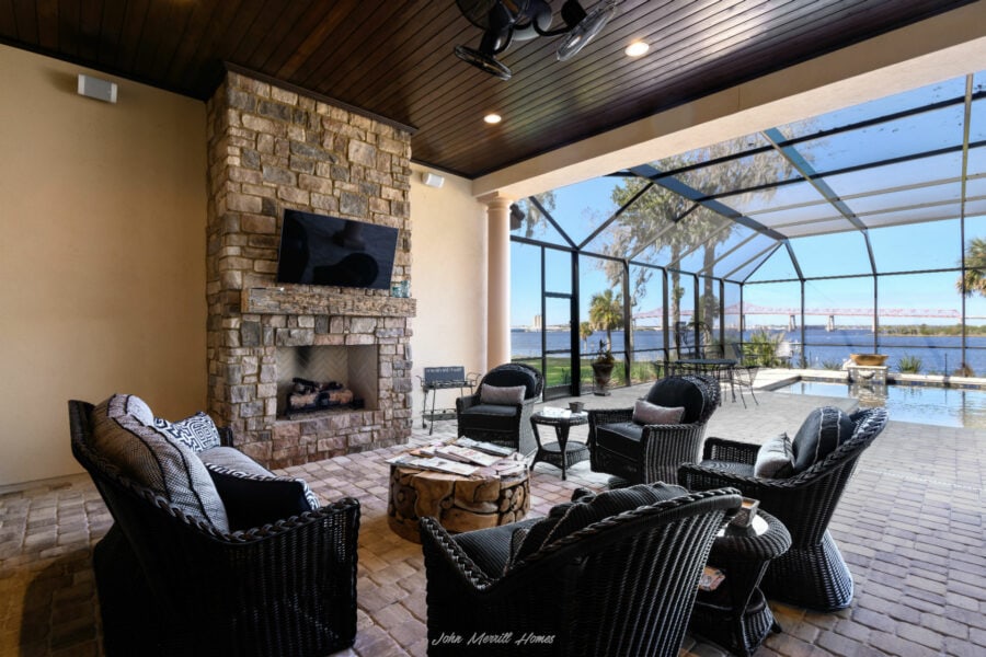 Modern enclosed patio with outdoor seating, stone fireplace, flat-screen TV, and view of water and bridge through screened windows.