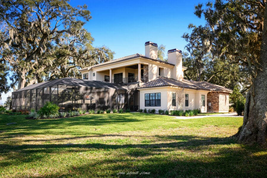 Luxury modern two-story home with a spacious backyard, covered patio, and lush landscaping, surrounded by mature moss-draped trees, under a clear blue sky.