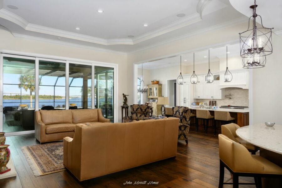 Modern living room with large glass sliding doors offering waterfront views, featuring tan leather sofas, patterned armchairs, and elegant decor, seamlessly connected to a bright, open kitchen and dining area.