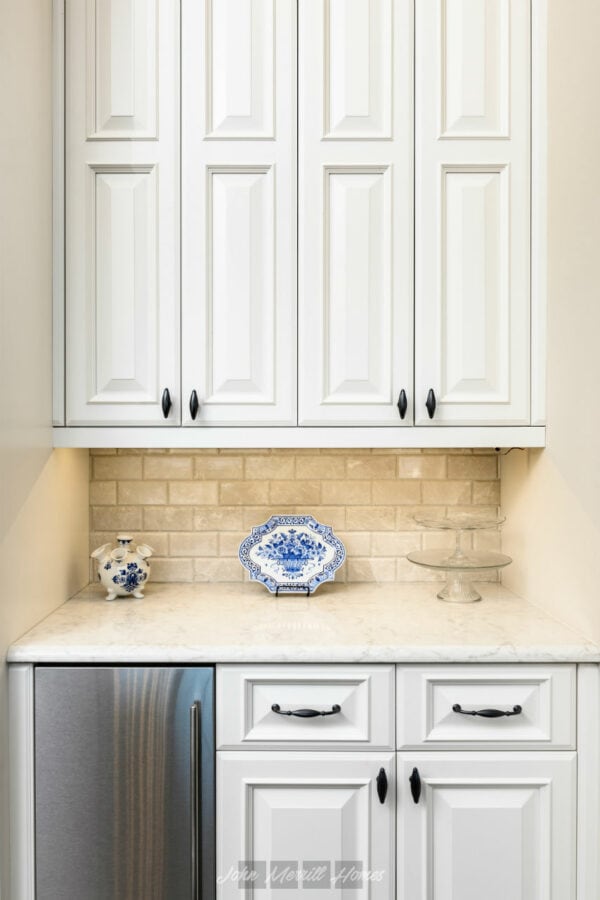 A kitchen cabinet with white paneled doors, black handles, and a beige brick backsplash, featuring decorative blue and white porcelain items on a marble countertop.