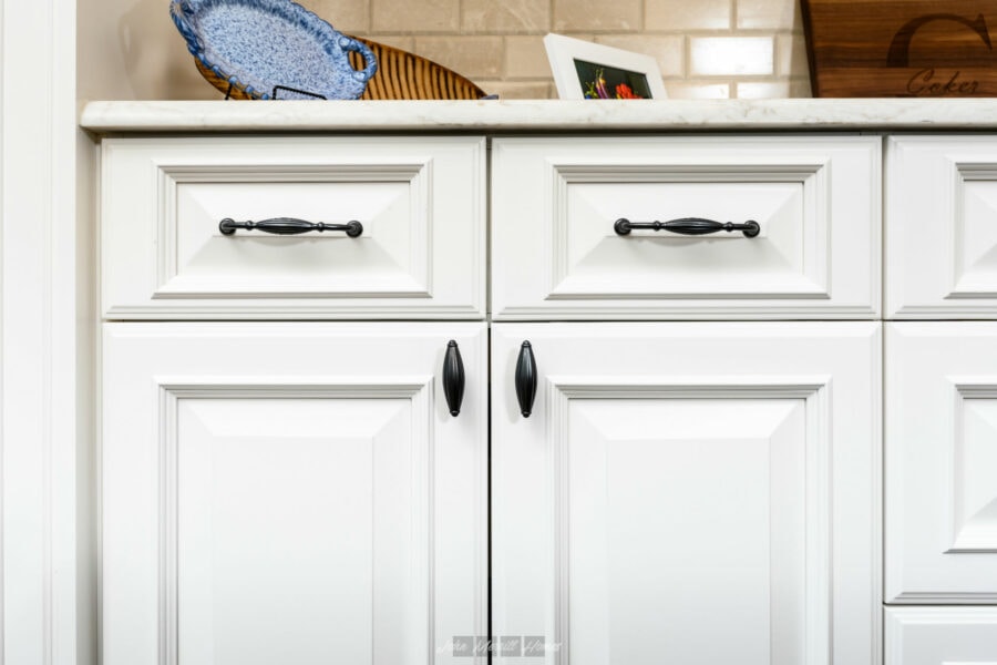 White kitchen cabinets with black handles and raised panel design, granite countertop, decorative items, and framed photo on beige brick wall background.