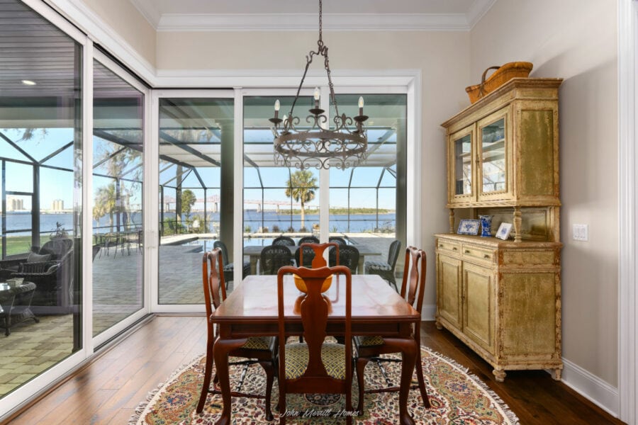 Elegant dining area with a wooden table, vintage chandelier, and a distressed yellow hutch overlooking a screened-in patio with water views. Bright, airy space blending rustic charm with modern comfort.