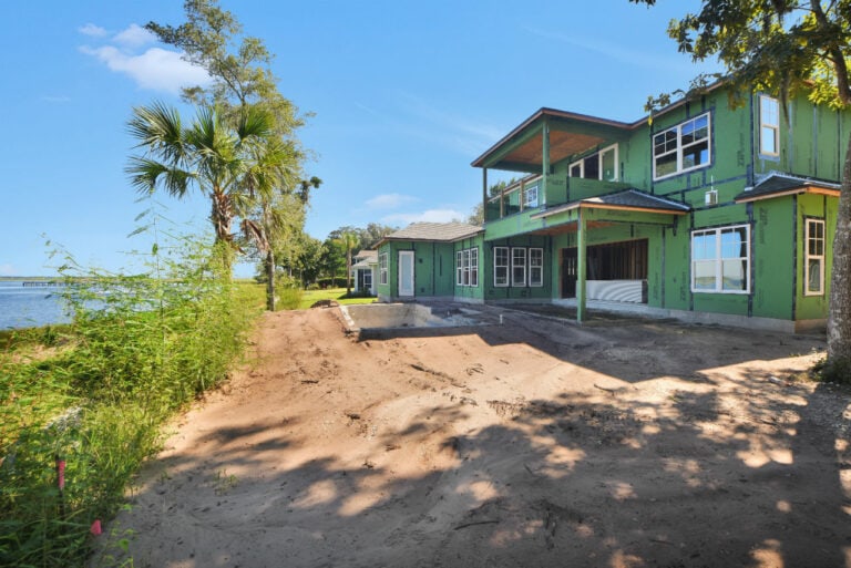 Newly under construction green house in a waterfront development area with palm trees and dirt driveway, featuring multiple windows and a large balcony, on a sunny day.