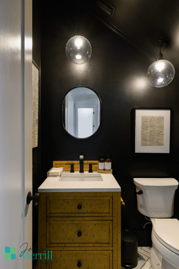 A contemporary bathroom featuring black walls, a wooden vanity with a white countertop, a round mirror, and modern lighting fixtures, creating a sleek and elegant space.