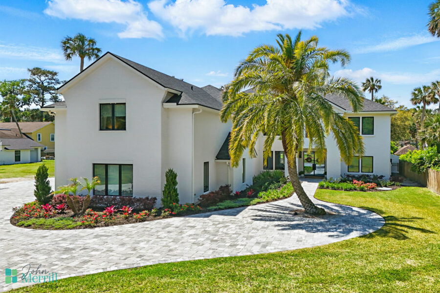 A spacious, contemporary home featuring a clean white exterior, large windows, and a well-maintained garden with colorful flowers and tall palm trees, set against a bright blue sky.
