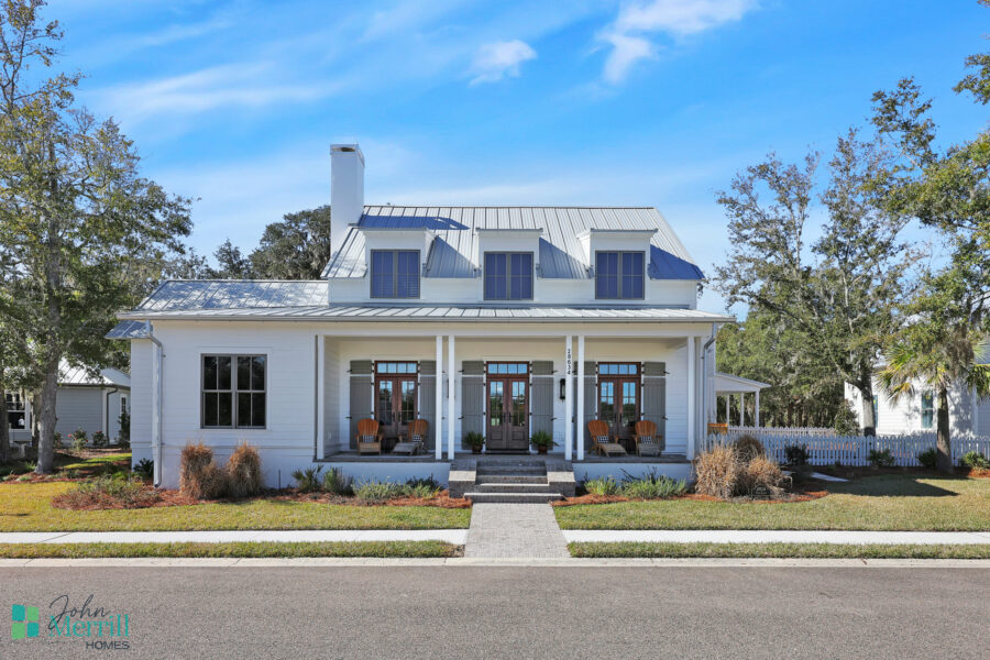 Beautiful white farmhouse with a spacious front porch, three front doors, and large windows, set against a clear blue sky with surrounding trees.