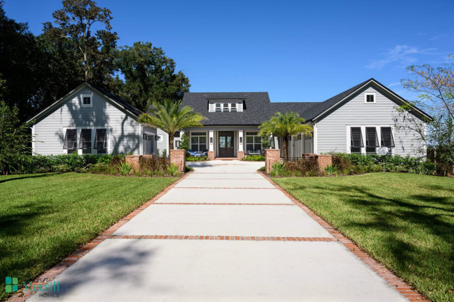 A spacious, contemporary white house featuring a welcoming front porch, surrounded by lush green lawns, palm trees, and well-maintained landscaping under a clear blue sky.