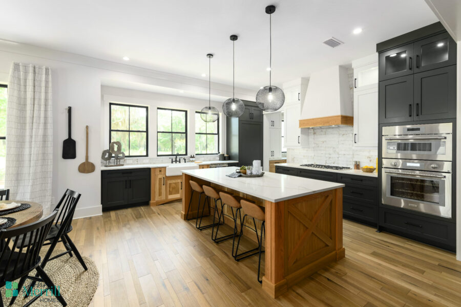 Bright, modern kitchen featuring black and wood cabinetry, a large island with seating, and stainless steel appliances, illuminated by natural light from large windows.