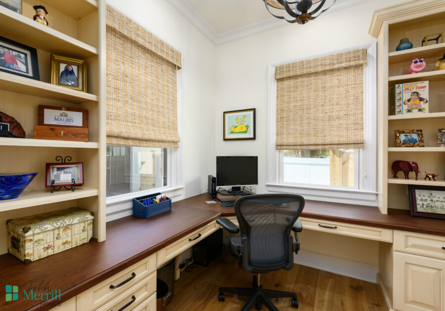 An organized home office featuring built-in shelves, a large wooden desk, and a comfortable ergonomic chair, illuminated by natural light from two windows with woven shades.