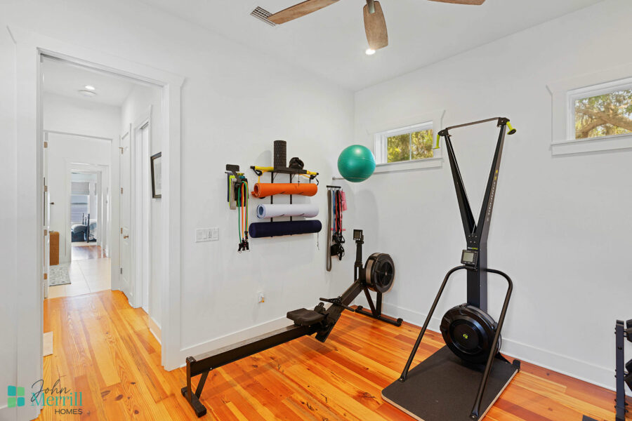 Home gym with various exercise equipment including a rowing machine, stability ball, resistance bands, and a wall-mounted storage rack in a well-lit room.