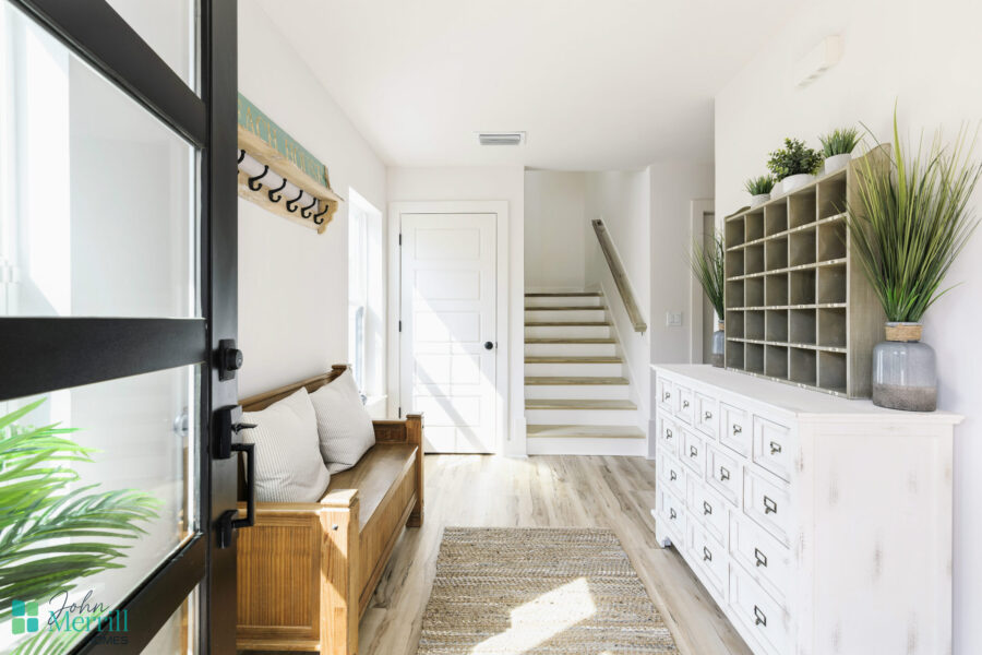 Bright and welcoming entryway featuring a white dresser with multiple drawers, decorative plants, and a wooden bench with cushions. Natural light illuminates the space, creating a cozy atmosphere.