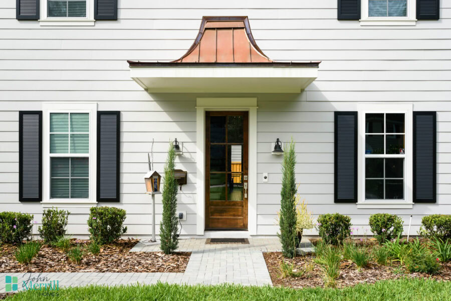Beautiful white house exterior with black shutters, a wooden front door, and a copper awning, surrounded by well-maintained landscaping and a paved walkway.
